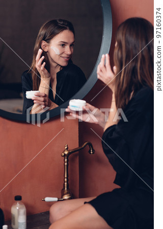 Young woman in front of the mirror putting cream on her face 97160374