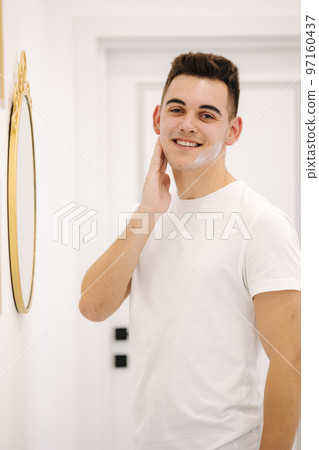 Handsome smiled man applies cream on the face. Male stand by mirror in beauty studio 97160437