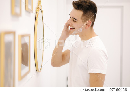 Handsome smiled man applies cream on the face. Male stand by mirror in beauty studio Handsome smiled man applies cream on the face. Male stand by mirror in beauty studio 97160438