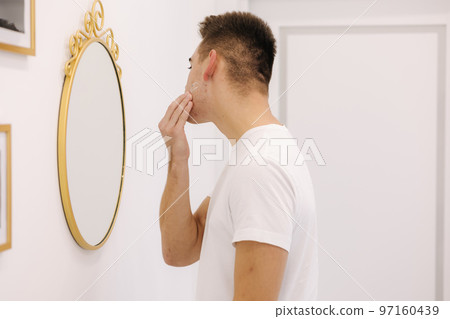 Handsome smiled man applies cream on the face. Male stand by mirror in beauty studio Handsome smiled man applies cream on the face. Male stand by mirror in beauty studio 97160439