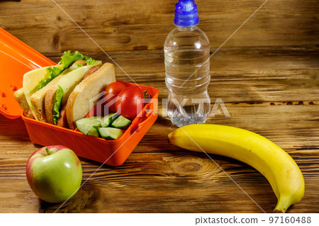 Bottle of water, apple, banana and lunch box with sandwiches and fresh vegetables on a wooden table 97160488