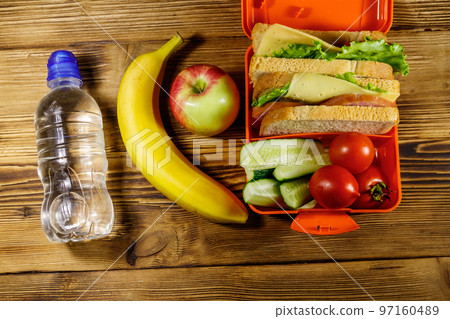 Bottle of water, apple, banana and lunch box with sandwiches and fresh vegetables on a wooden table. Top view Bottle of water, apple, banana and lunch box with sandwiches and fresh vegetables on a wooden table. Top view 97160489