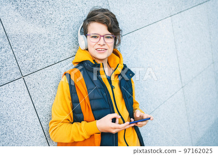 Child caucasian teen boy yellow hoodie with wireless headphones looks into camera outdoors using smartphone. communication on the go. Candid portrait Blogging vlogging surfing Music Video call. Gaming 97162025
