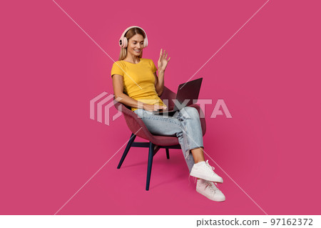 Happy mature caucasian blonde woman in casual and headphones sits on armchair, waving hand at laptop 97162372