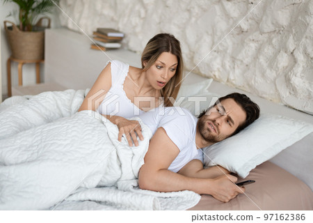 Shocked woman seeing her husband messaging to his lover on cellphone while lying in bed near him 97162386