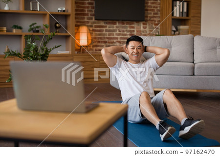 Smiling asian mature man working on abs, watching fitness tutorial on laptop and exercising on fitness mat at home 97162470