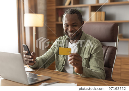Glad mature black man uses credit card, laptop and phone to check finances at workplace Glad mature black man uses credit card, laptop and phone to check finances at workplace 97162532