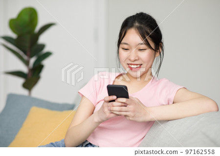 Excited korean lady sitting on sofa and using smartphone, chatting with friends online, checking social media Excited korean lady sitting on sofa and using smartphone, chatting with friends online, checking social media 97162558