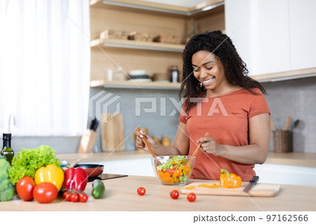 Vegetarian lifestyle. Cheerful pretty millennial black lady in red t-shirt make salad, prepares lunch for family 97162566