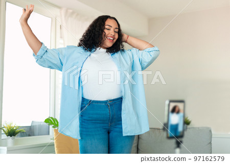 African american plus size female influencer recording video on smartphone, dancing on camera at home 97162579