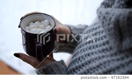 Close-up of young woman in grey knitted sweater drinking tea or hot chocolate with marshmallows on winter holidays Close-up of young woman in grey knitted sweater drinking tea or hot chocolate with marshmallows on winter holidays 97162894