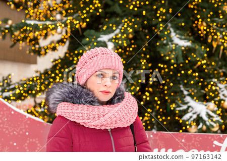 A young woman in a pink hat and scarf in a snowy city in winter at the Christmas tree. Looks to the future with hope. Close-up. Space for text. Copy space 97163142