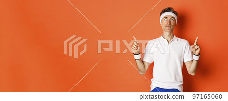 Portrait of handsome middle-aged male athlete, wearing sports clothing, looking and pointing fingers up at promo banner, standing against orange background 97165060