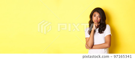 Portrait of unamused and bored african-american teenage girl, looking reluctant at camera, standing over yellow background Portrait of unamused and bored african-american teenage girl, looking reluctant at camera, standing over yellow background 97165341