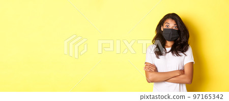 Concept of coronavirus, pandemic and lifestyle. Image of surprised african-american girl in face mask, looking amazed at something cool, standing over yellow background 97165342