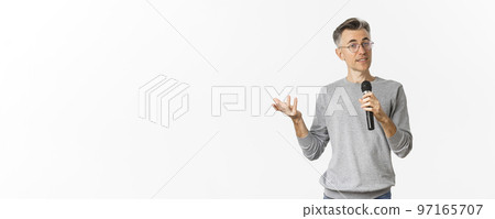 Portrait of handsome middle-aged man in glasses and gray sweater, making speech, talking with microphone and explaining something, standing over white background 97165707