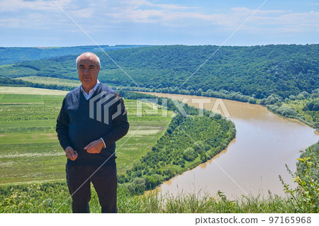 An elderly cheerful man is a pensioner standing on a hill near the river. An elderly cheerful man is a pensioner standing on a hill near the river. 97165968