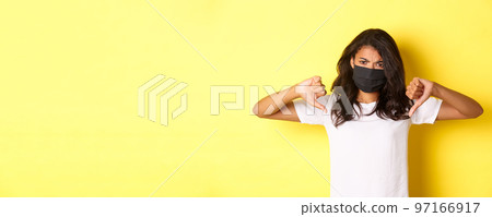 Concept of covid-19, social distancing and lifestyle. Image of upset african-american female protester, wearing black face mask, showing thumbs-down and frowning disappointed, yellow background 97166917