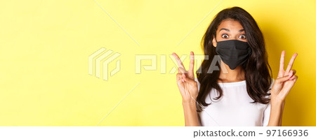 Concept of coronavirus, pandemic and lifestyle. Close-up of silly african-ameircan girl in black face mask, showing peace signs and standing over yellow background Concept of coronavirus, pandemic and lifestyle. Close-up of silly african-ameircan girl in black face mask, showing peace signs and standing over yellow background 97166936