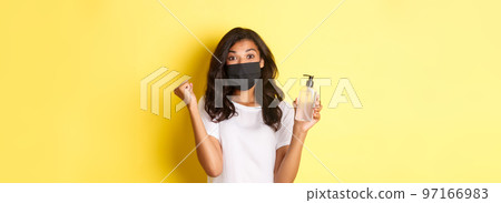 Concept of covid-19, social distancing and lifestyle. Image of happy african-american woman in face mask, feeling happy about founding good hand sanitizer, rejoicing over yellow background Concept of covid-19, social distancing and lifestyle. Image of happy african-american woman in face mask, feeling happy about founding good hand sanitizer, rejoicing over yellow background 97166983