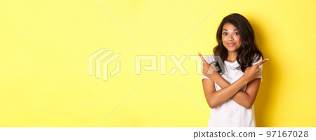 Portrait of cute indecisive african american girl, pointing fingers sideways and shrugging, asking advice with choices, standing over yellow background 97167028