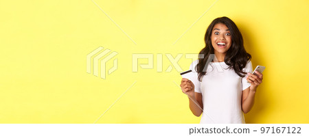Portrait of amazed african-american girl, holding smartphone and credit card, standing over yellow background Portrait of amazed african-american girl, holding smartphone and credit card, standing over yellow background 97167122