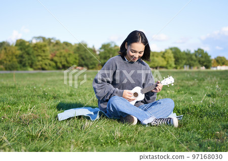Singing asian girl playing ukulele on grass, sitting on blanket in park, relaxing outdoors on sunny day 97168030