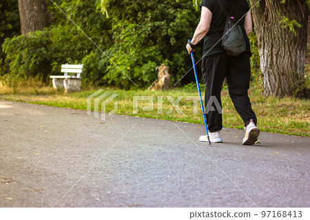 Nordic walking with sticks, a type of physical activity for retirees. An elderly middle-aged woman does Scandinavian walking outside in a summer park on nature. Sporty active faceless woman back view. Nordic walking with sticks, a type of physical activity for retirees. An elderly middle-aged woman does Scandinavian walking outside in a summer park on nature. Sporty active faceless woman back view. 97168413