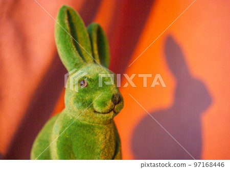 Green cute funny Easter bunny with long ears on the orange sunny background in springtime. Easter holiday in April. Toy decorative dreamy hare - a symbol of Easter. Smiling rabbit portrait close-up Green cute funny Easter bunny with long ears on the orange sunny background in springtime. Easter holiday in April. Toy decorative dreamy hare - a symbol of Easter. Smiling rabbit portrait close-up 97168446
