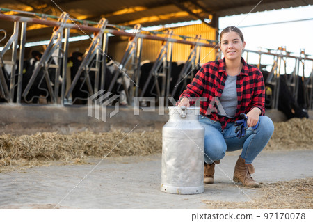Smiling girl dairy farm worker posing with milk can in cowshed Smiling girl dairy farm worker posing with milk can in cowshed 97170078