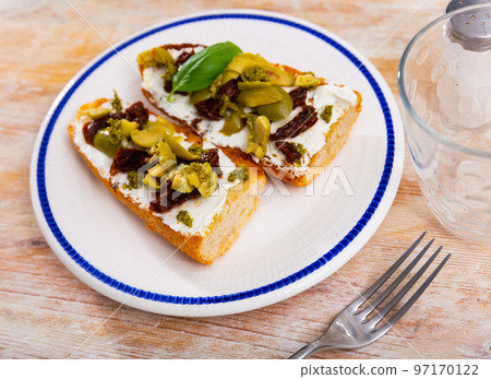Tasty sandwich with cheese, sun-dried tomatoes, pesto sauce and olives on the white plate with blue line on the tablecloth Tasty sandwich with cheese, sun-dried tomatoes, pesto sauce and olives on the white plate with blue line on the tablecloth 97170122