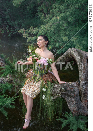 Blooming gorgeous lady in dress of flowers sitting on tree over water in the morning fog 97175658