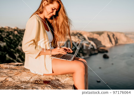 Digital nomad, Business woman working on laptop by the sea. Pretty lady typing on computer by the sea at sunset, makes a business transaction online from a distance. Freelance, remote work on vacation 97176064