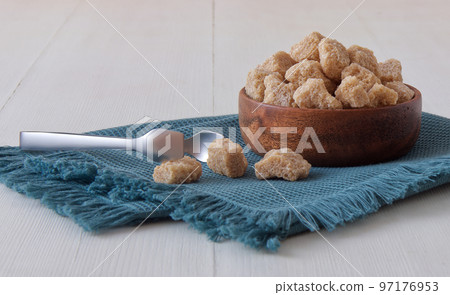 Cube-shaped cane sugar in wooden bowl on a white wooden table with silver sugar tongs. Low angle view, no people. Cube-shaped cane sugar in wooden bowl on a white wooden table with silver sugar tongs. Low angle view, no people. 97176953