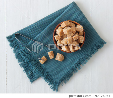 Cube-shaped cane sugar in wooden bowl on a turquoise napkin with silver sugar tongs. Flat lay, top down view, no people. 97176954