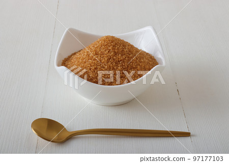 Granulated cane sugar in porcelain bowl on a white wooden table with a golden spoon. Low angle view, no people. 97177103