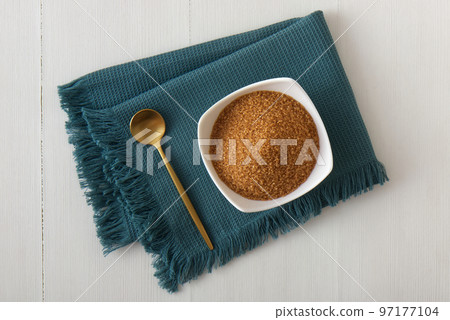 Granulated cane sugar in porcelain bowl on a turquoise napkin with a golden spoon. High angle view, white wooden table, no people. 97177104