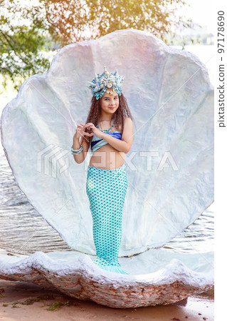 A little girl in a mermaid costume is standing outdoors, in a large seashell 97178690