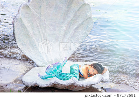 A pretty little girl in a turquoise mermaid costume sleeping in a large sea shell 97178691