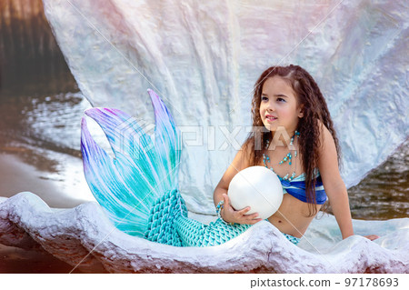 A beautiful little girl in a mermaid costume sits in a large sea shell with big pearl A beautiful little girl in a mermaid costume sits in a large sea shell with big pearl 97178693