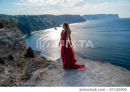 A girl with flowing hair in a long red dress stands on a rock above the sea. The stone can be seen in the sea. Sunny path to the sea from the sun. A girl with flowing hair in a long red dress stands on a rock above the sea. The stone can be seen in the sea. Sunny path to the sea from the sun. 97180906