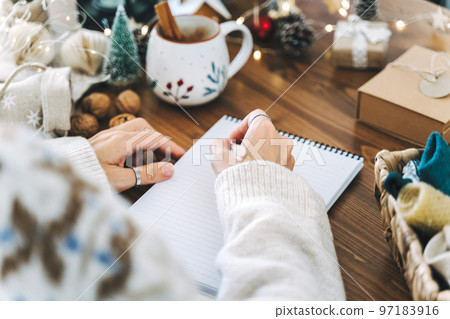 Woman hand holding pen on notepad at home on winter holidays xmas. Goals plans make to do and wish list for new year christmas concept, girl writing in notebook. Christmas decoration, gift boxes Woman hand holding pen on notepad at home on winter holidays xmas. Goals plans make to do and wish list for new year christmas concept, girl writing in notebook. Christmas decoration, gift boxes 97183916