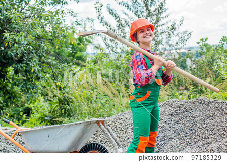 teen girl takes out rubble from wheelbarrow. kid with shovel loading crushed stones. Laying the foundation, building project. girl with wheelbarrow of rubble. kid working on construction site teen girl takes out rubble from wheelbarrow. kid with shovel loading crushed stones. Laying the foundation, building project. girl with wheelbarrow of rubble. kid working on construction site 97185329