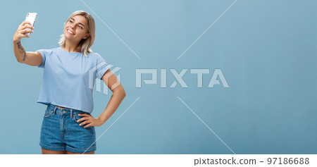 Stylish cute female gym manager taking selfie with new smartphone posing with broad pleased smile and holding hand on hip making post for followers standing carefree and relaxed over blue wall Stylish cute female gym manager taking selfie with new smartphone posing with broad pleased smile and holding hand on hip making post for followers standing carefree and relaxed over blue wall 97186688