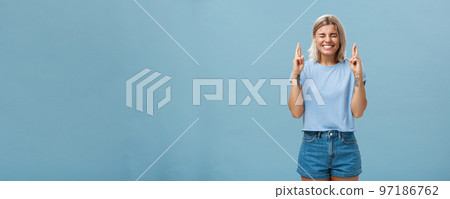 Optimistic faithful good-looking young woman with blond hair and tattoos smiling joyfully crossing fingers for good luck waiting for dream come true making wish while standing over blue background Optimistic faithful good-looking young woman with blond hair and tattoos smiling joyfully crossing fingers for good luck waiting for dream come true making wish while standing over blue background 97186762