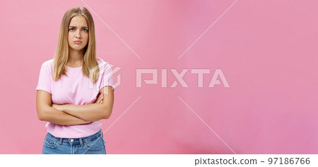 Sad envy and disappointed gloomy cute young woman with fair hair in casual t-shirt crossing arms against chest in upset gesture pursing lips and frowning looking concerned and sad over pink wall 97186766