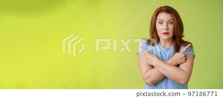 Help me choose. Worried unsure doubtful cute redhead middle-aged silly woman asking advice cross arms body pointing sideways left right look concerned camera make difficult decision green background 97186771