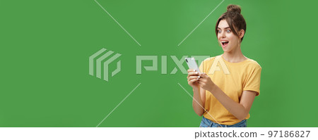 Woman reading surprising satisfying message in smartphone opening mouth from excitement, smiling amazed looking astonished at cellphone screen posing against green background in casual yellow t-shirt 97186827