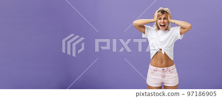Portrait of panicking concerned and depressed young european woman with blond hair tattoos and athletic body yelling out loud holding hands on head being concerned and shook over purple background Portrait of panicking concerned and depressed young european woman with blond hair tattoos and athletic body yelling out loud holding hands on head being concerned and shook over purple background 97186905