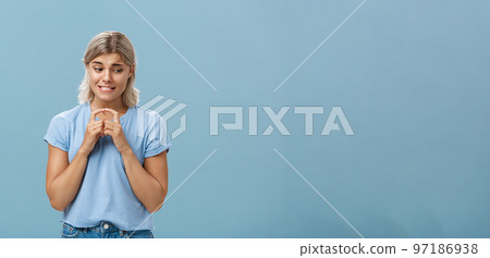 Studio shot of insecure and timid silly girlfriend with blond hair clenching teeth looking shy down and steepling index fingers unconfident trying ask out boy she likes over blue background Studio shot of insecure and timid silly girlfriend with blond hair clenching teeth looking shy down and steepling index fingers unconfident trying ask out boy she likes over blue background 97186938